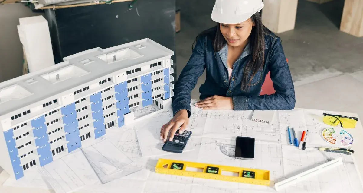 Architektka w kasku analizuje projekt budynku, używając kalkulatora. To może być związane z bms co to jest za system.