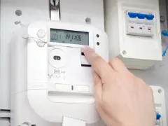 Dłoń naciska przycisk na ekranie inteligentnego licznika energii elektrycznej. Obok widoczny jest wyłącznik.