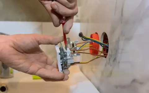 Instalator przykręca gniazdko, pokazując, jak podłączyć gniazdko. Widać przewody elektryczne i narzędzia.