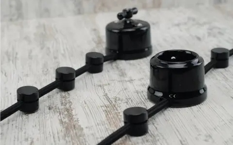 Stylowa instalacja elektryczna natynkowa z czarnymi ceramicznymi elementami: gniazdkiem i przełącznikiem, na drewnianym tle.