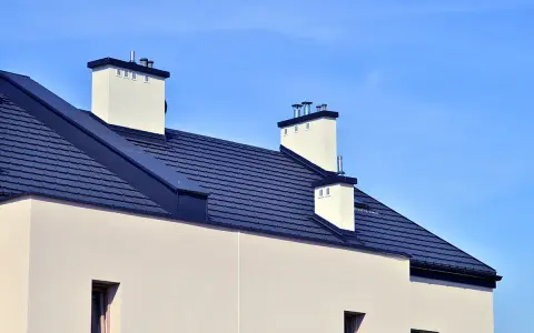 Nowy dom z ciemnym, dachówkowym dachem bezokapowym i białymi kominami, pod błękitnym niebem.
