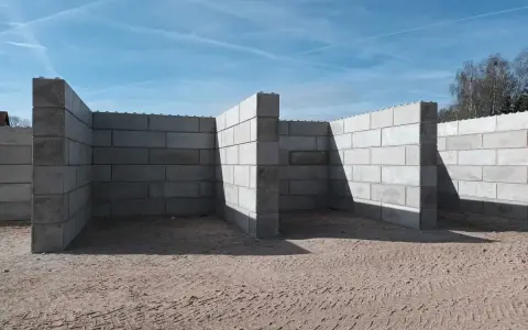 Budowa muru oporowego z bloczków betonowych. Konstrukcja w kształcie litery L, z widocznymi cieniami na piasku.