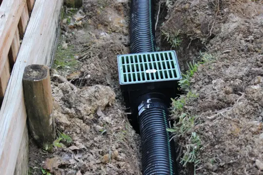 Instalacja drenażu przy drewnianym płocie. W wykopie widać czarną rurę i zieloną kratkę odpływową.