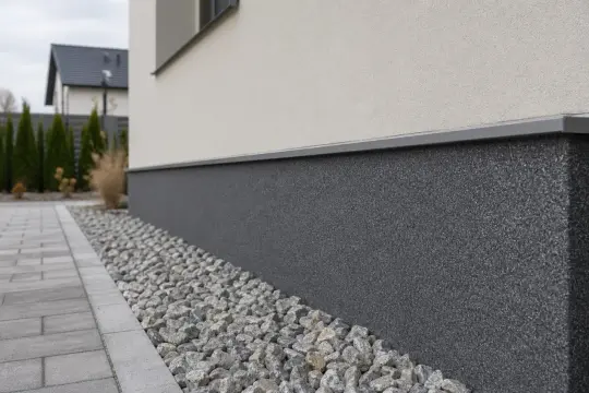 Nowoczesny cokół domu z ciemnoszarą fakturą, otoczony kamieniami i chodnikiem z kostki brukowej.