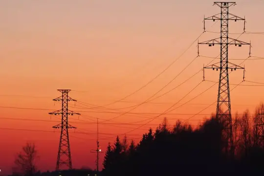 Wieczorny krajobraz z liniami energetycznymi. Wysokie słupy przesyłowe na tle pomarańczowego nieba, symbolizujące dodatek energetyczny dla domów.