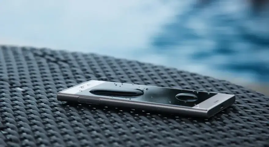 Smartfon z kroplami wody na ekranie, leżący na plecionej powierzchni. W tle widać basen. IP68 co oznacza? Ochronę przed wodą i pyłem.