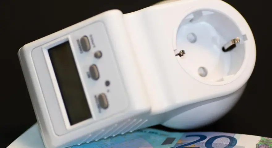 Biały miernik zużycia prądu z wyświetlaczem i przyciskami, leżący na banknocie 20 euro.