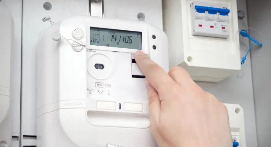 Dłoń naciska przycisk na ekranie inteligentnego licznika energii elektrycznej. Obok widoczny jest wyłącznik.