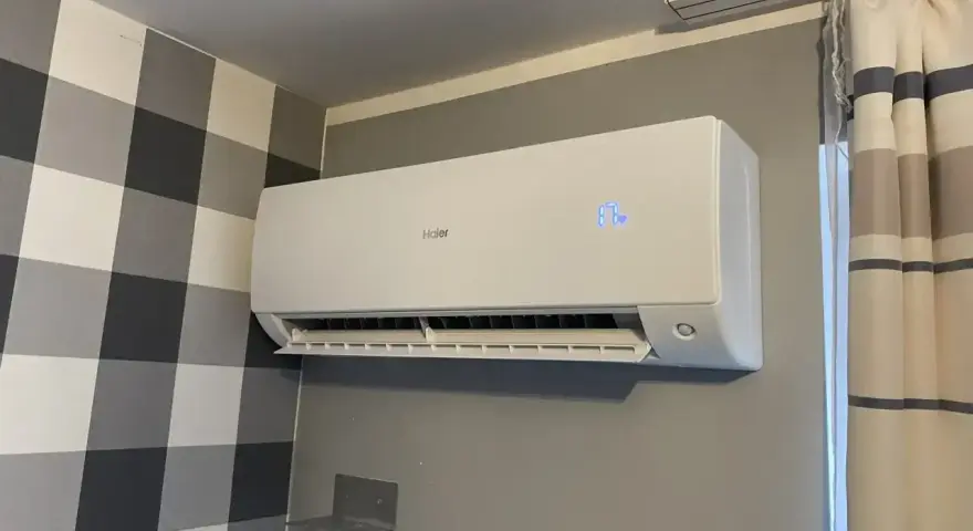 Biała klimatyzacja Haier zamontowana na szarej ścianie, obok wzorzystej tapety i zasłon.