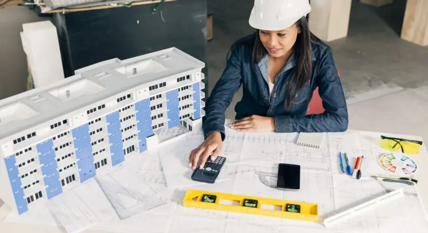Architektka w kasku analizuje projekt budynku, używając kalkulatora. To może być związane z bms co to jest za system.