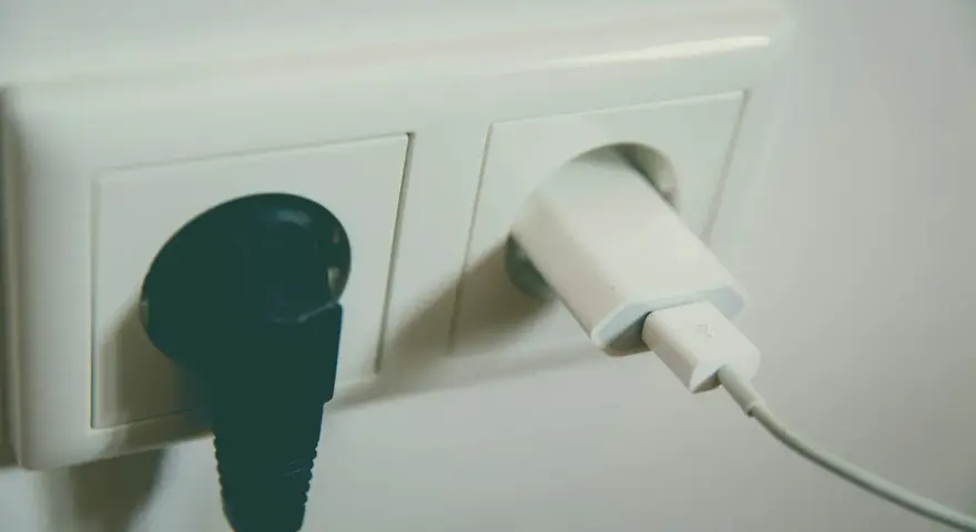 Dwa gniazdka elektryczne. W jednym czarna wtyczka, w drugim biała ładowarka. Czekają na napięcie w gniazdku.